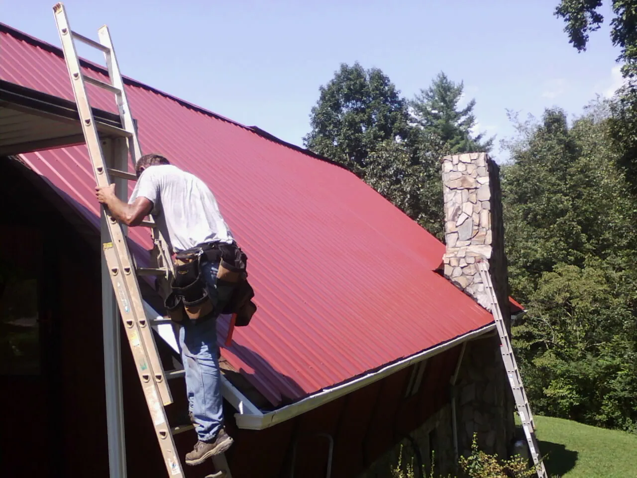 Roofer on a ladder beside a red metal roof and stone chimney — Soffit & Fascia Repair by Roofing Brothers in Alexander